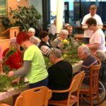 Več oseb sedi za mizo in skupaj izdeluje šopke iz vejic in cvetja.