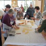 Skupina ljudi stoji okoli mize in skupaj pripravlja testo ter izdeluje rezance s pomočjo kuhinjskih pripomočkov.