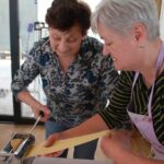 Dve osebi uporabljata strojček za valjanje testa in skupaj oblikujeta trakove za rezance.