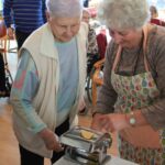 Dve starejši osebi sodelujeta pri pripravi testa in ga valjata skozi strojček, medtem ko drugi opazujejo v ozadju.