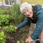 Oseba se sklanja nad gredo in se dotika rastline na zunanjem vrtu ob stavbi.
