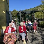 Več oseb sedi na vozičkih in klopeh na sončnem dvorišču ob stavbi.