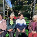 Štiri osebe sedijo na zeleni klopi na prostem pod pergolo.