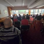 Starejši stanovalci sedijo v prostoru in gledajo projekcijo filma na velikem platnu; nekateri so v invalidskih vozičkih.