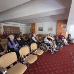 Pogled s strani na občinstvo, ki mirno spremlja film; v ospredju so prazni stoli, zadaj pa gledalci.