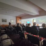 Temnejši prizor med projekcijo, kjer gledalci sedijo obrnjeni proti platnu in pozorno spremljajo vsebino.