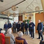 Skupina moških stoji v polkrogu in poje pred starejšimi stanovalci; prostor je okrašen z naravnimi velikonočnimi dekoracijami.