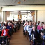 Številni starejši stanovalci sedijo na stolih in poslušajo nastop; nekateri uporabljajo hodulje ali invalidske vozičke.