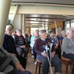 Pogled iz bližine na občinstvo starejših, ki pozorno spremljajo dogajanje; v ospredju je ženska z berglami.