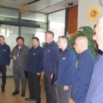 Pevska skupina moških med nastopom, oblečena v enotna temna oblačila, stoji v prostoru doma za starejše.