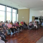 Skupina oseb sedi na stolih in invalidskih vozičkih ter posluša osebi, ki stojita pred njimi.