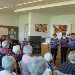 Skupina oseb stoji v vrsti in poje iz map, pred njimi pa sedijo osebe, ki jih poslušajo.