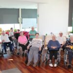 Osebe sedijo v prostoru na stolih in invalidskih vozičkih ter gledajo proti dogajanju izven kadra.