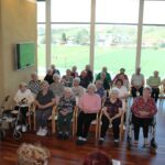 Več oseb sedi v več vrstah na stolih in invalidskih vozičkih ter je obrnjenih proti sprednjemu delu prostora.