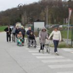 Skupina oseb hodi po cesti in prečka prehod za pešce, pri čemer nekatere osebe uporabljajo pripomočke za hojo ali vozičke.