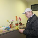 Oseba stoji ob polici in v roki drži umetno rožo, medtem ko si ogleduje razstavljene predmete.