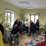 Več oseb se giblje po prostoru in si ogleduje razstavljene okraske na mizah in vejah.