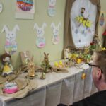 Oseba stoji pred razstavno mizo in opazuje različne ročno izdelane okraske.