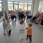 Otroci stojijo na sredini prostora in se držijo za roke, okoli njih pa sedijo osebe, ki jih opazujejo.