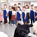 Otroci stojijo v vrsti in nastopajo pred sedečimi osebami v prostoru.