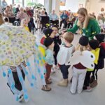 Otroci v kostumih stojijo v krogu, ena oseba jim drži mikrofon, v ozadju pa sedijo osebe, ki jih opazujejo.