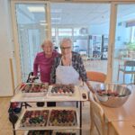 Dve starejši gospe stojita za vozičkom, polnim ročno pobarvanih pirhov, in se nasmejani predstavita ob svojem delu.