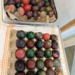 Pladenj z raznobarvnimi pirhi, okrašenimi z različnimi vzorci in naravnimi barvami, razporejenimi v kartonski embalaži.