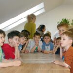 Otroci radovedno sedijo za mizo in z zanimanjem opazujejo priprave na ustvarjalno delavnico.