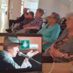 Več oseb sedi v prostoru in gleda prenos nastopa na prenosnem računalniku, postavljenem na mizi.