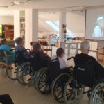Več oseb sedi na stolih in invalidskih vozičkih ter gleda projekcijo nastopa na platnu v skupnem prostoru.