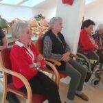 Več oseb sedi v vrsti na stolih in invalidskih vozičkih ter spremlja dogajanje izven kadra.