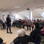 Stanovalci z zanimanjem spremljajo nastop, kjer gostja z mikrofonom ustvarja prijetno in sproščeno vzdušje.