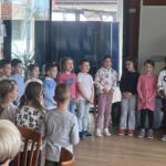 Skupina otrok stoji v vrsti in poje, ob njih stoji odrasla oseba, ki jih vodi. V ozadju sedijo starejši poslušalci.