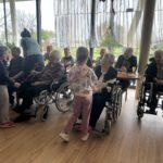 Otroci se družijo s starejšimi osebami; ena deklica podarja rožo starejši gospe na vozičku, ostali opazujejo in uživajo v trenutku.