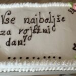 Na mizi je velika torta z napisom »Vse najboljše za rojstni dan!«, okrašena s cvetovi iz sladkorne mase.