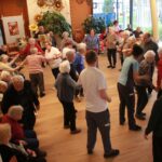 Večja skupina starejših oseb stoji v krogu in pleše oziroma se giba ob glasbi, nekateri sedijo ob strani in opazujejo.
