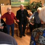 Starejši moški in ženski se držijo za roke v krogu ter sodelujejo v skupinskem plesu ali telovadbi.