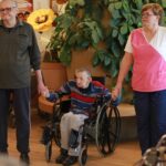 Starejši moški na invalidskem vozičku sodeluje v krogu, medtem ko se z obeh strani drži za roke z drugimi udeleženci aktivnosti.