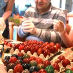 Moški z zaščitnimi rokavicami sedi za mizo in sodeluje pri barvanju pirhov, pred njim so pladnji z že pobarvanimi jajci v različnih odtenkih.