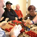 Več starejših oseb sedi okoli mize in skupaj barva pirhe; na mizi so pladnji z jajci in steklenica barve ali tekočine.