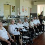 Več oseb sedi ob steni v skupnem prostoru, pred njimi stoji deček z harmoniko in igra.