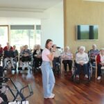 Oseba poje v mikrofon na sredini prostora, okoli nje sedijo osebe na stolih in invalidskih vozičkih.