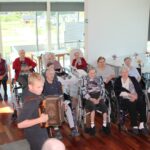 Deček z harmoniko stoji pred skupino sedečih oseb v svetlem prostoru z velikimi okni.