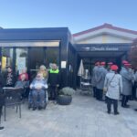 Skupina oseb stoji in sedi pred vhodom v stavbo, nekatere osebe so na vozičkih, ob strani stoji skupina oseb v enotnih oblačilih.