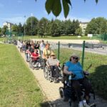 Skupina stanovalcev in spremljevalcev se po poti sprehaja ob ograjenem športnem igrišču.