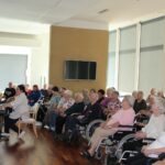 Pevski zbor in pianistka nastopajo pred zbranimi stanovalci v domu za starejše.