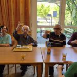 Skupina stanovalcev sedi za mizami in z dvignjenimi kozarci nazdravlja ob rojstnodnevni torti.
