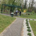 Dve osebi urejata zemljo ob pergoli na zelenici ob poti.