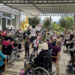 Stanovalci in otroci sodelujejo pri skupinski telovadbi na zunanji terasi doma za starejše.