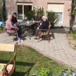 Skupina oseb sedi za mizo na zunanjem dvorišču ob stavbi.